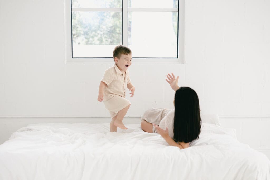 Candid moment of mom laying on the bed while her toddler laughs and walks around during a Tampa family maternity session.