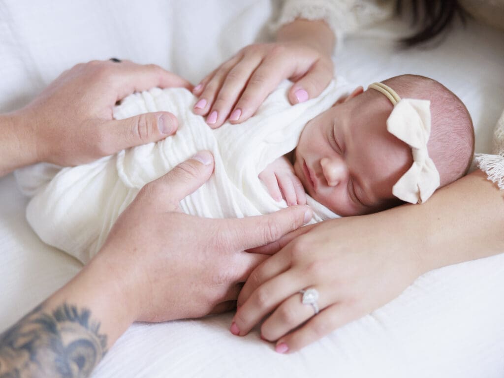 Sweet newborn baby girl swaddled in a cream colored wrap with a bow headband surrounded by parents hands in Tampa newborn photography session