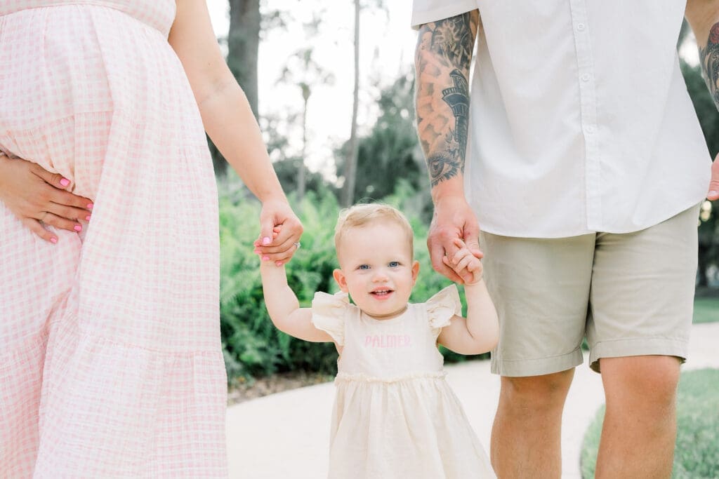 Toddler holding parents hands with a natural pregnancy after an IVF pregnancy in Sims Park for a Tampa Maternity Photography Session.
