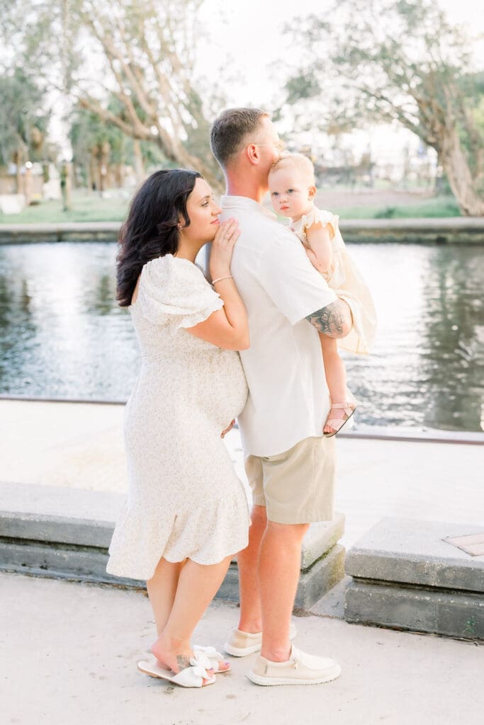Mom experiencing a natural pregnancy after an IVF pregnancy with husband holding toddler looking at sunset in Sims Park for a Tampa Maternity Photography Session.