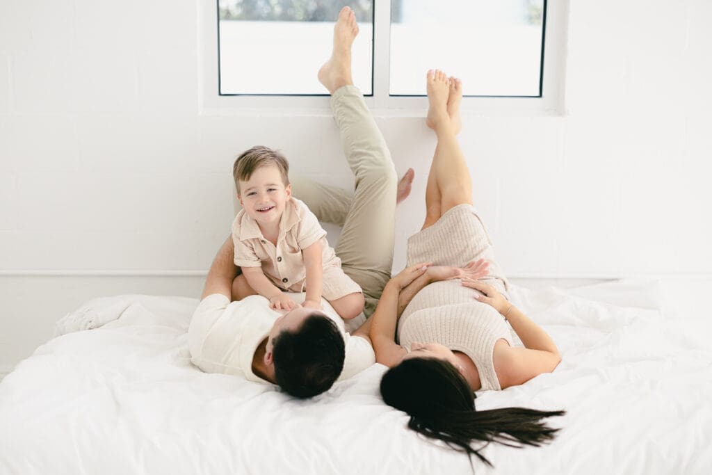 Parents laying together on the bed during a Tampa maternity session while their toddler sits on dad and smiles at the camera.